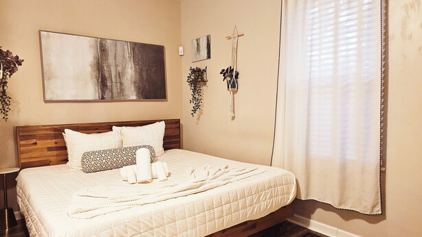 A bedroom with a wooden headboard, a large abstract painting, a macrame wall hanging, and a window with sheer curtains.