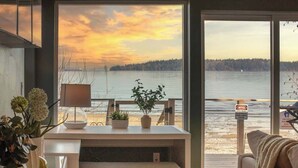 Een moderne woonkamer met een groot raam met uitzicht op een meer, een kleine tafel met een lamp en planten, en een bank.