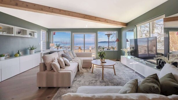 Ein großzügiger Wohnraum mit großem Fernseher, bequemen Sitzgelegenheiten und Blick auf den Strand durch große Fenster.