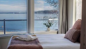 Een slaapkamer met een groot raam met uitzicht op een wateroppervlak en een bebost gebied.