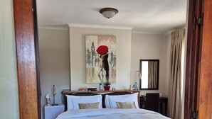 Un dormitorio con piso de madera, una cama con ropa de cama blanca, un cuadro en la pared y una mesita de noche con una lámpara.