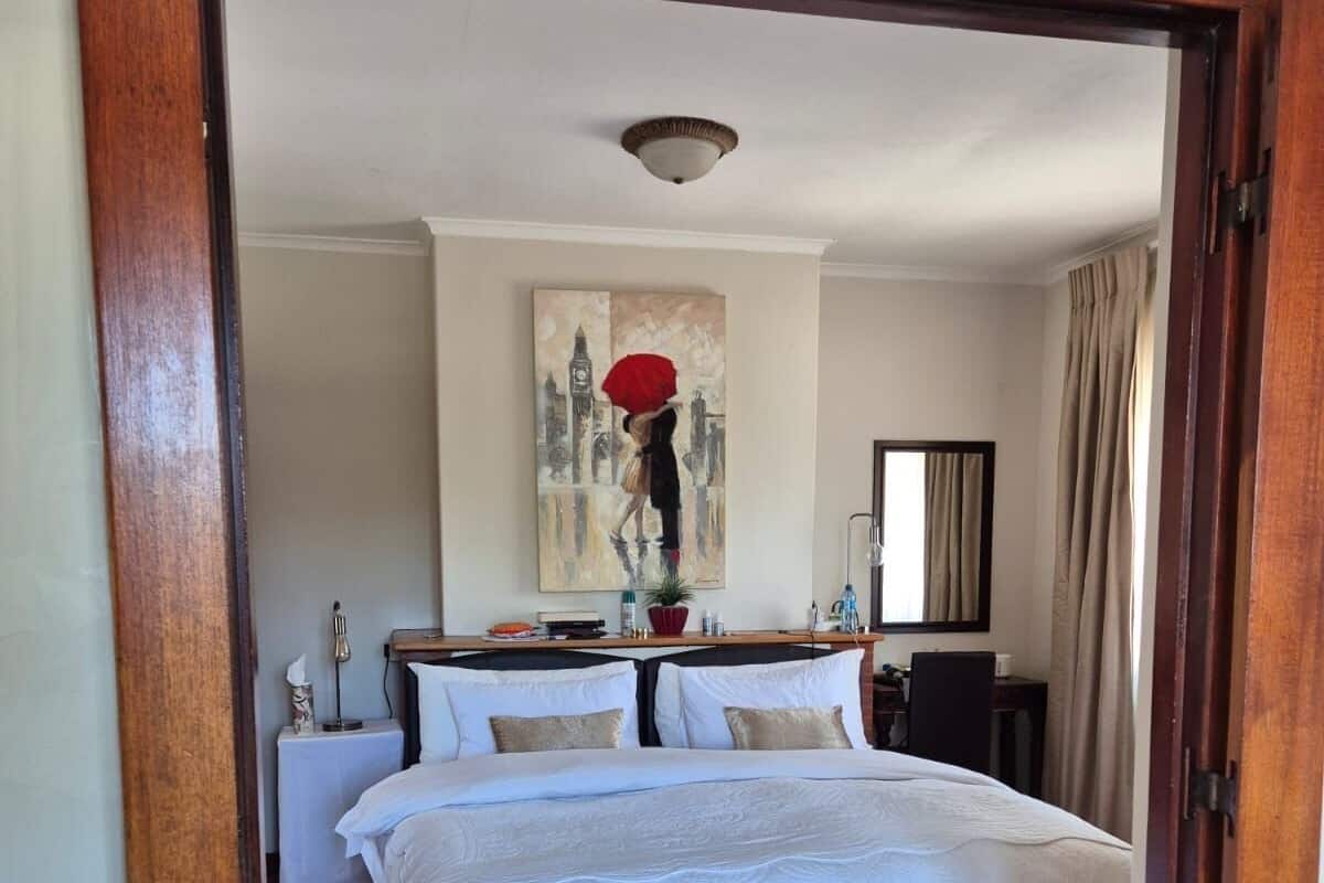 A bedroom with a wooden floor, a bed with white bedding, a painting on the wall, and a nightstand with a lamp.
