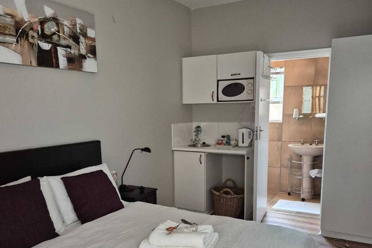 A neatly made bed with white linens and a folded towel, a white cabinet with a microwave, a small kitchenette, and a bathroom visible through an open door.