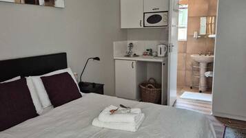 A neatly made bed with white linens and a folded towel, a white cabinet with a microwave, a small kitchenette, and a bathroom visible through an open door.