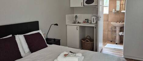 A neatly made bed with white linens and a folded towel, a white cabinet with a microwave, a small kitchenette, and a bathroom visible through an open door.