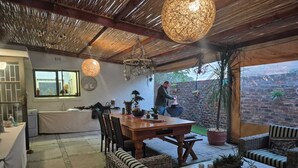 Un patio al aire libre con una mesa de madera y sillas de mimbre, un muro de ladrillo y una persona asando.