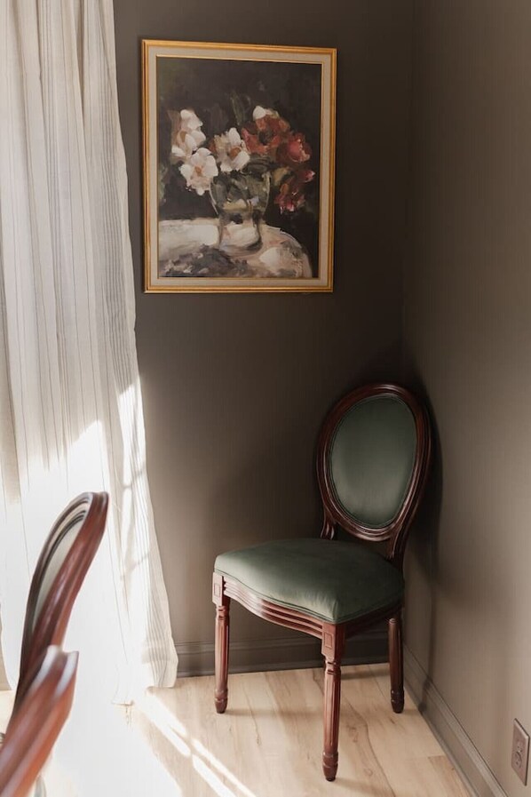 Una habitación con una pared oscura, un cuadro enmarcado de flores, una butaca tapizada de verde y una silla de madera.