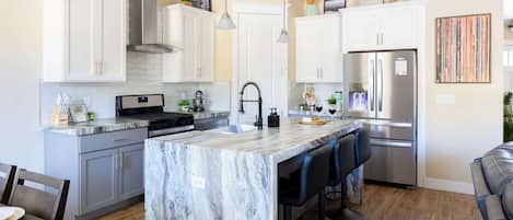Una cocina moderna con armarios blancos, una isla de mármol, electrodomésticos de acero inoxidable y un comedor con mesa y sillas de madera.