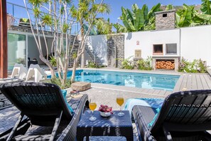 Zona de piscina con sillones de descanso, una mesa con bebidas y una pared de piedra con una parrilla.