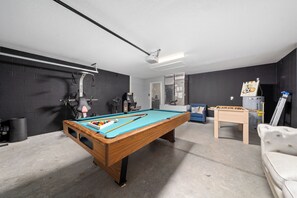 Una mesa de billar con cubierta de fieltro azul, una mesa de fútbol de mesa y un sofá blanco en una sala con paredes de ladrillo oscuro y techo blanco.