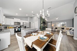 Una cocina moderna con armarios blancos, un comedor con mesa de madera dispuesta para cuatro personas y una sala de estar con sofá y televisor.