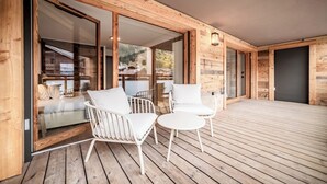 A wooden deck with two white chairs and a small table, large sliding glass doors leading to a bedroom with a bed and a view of a mountain.