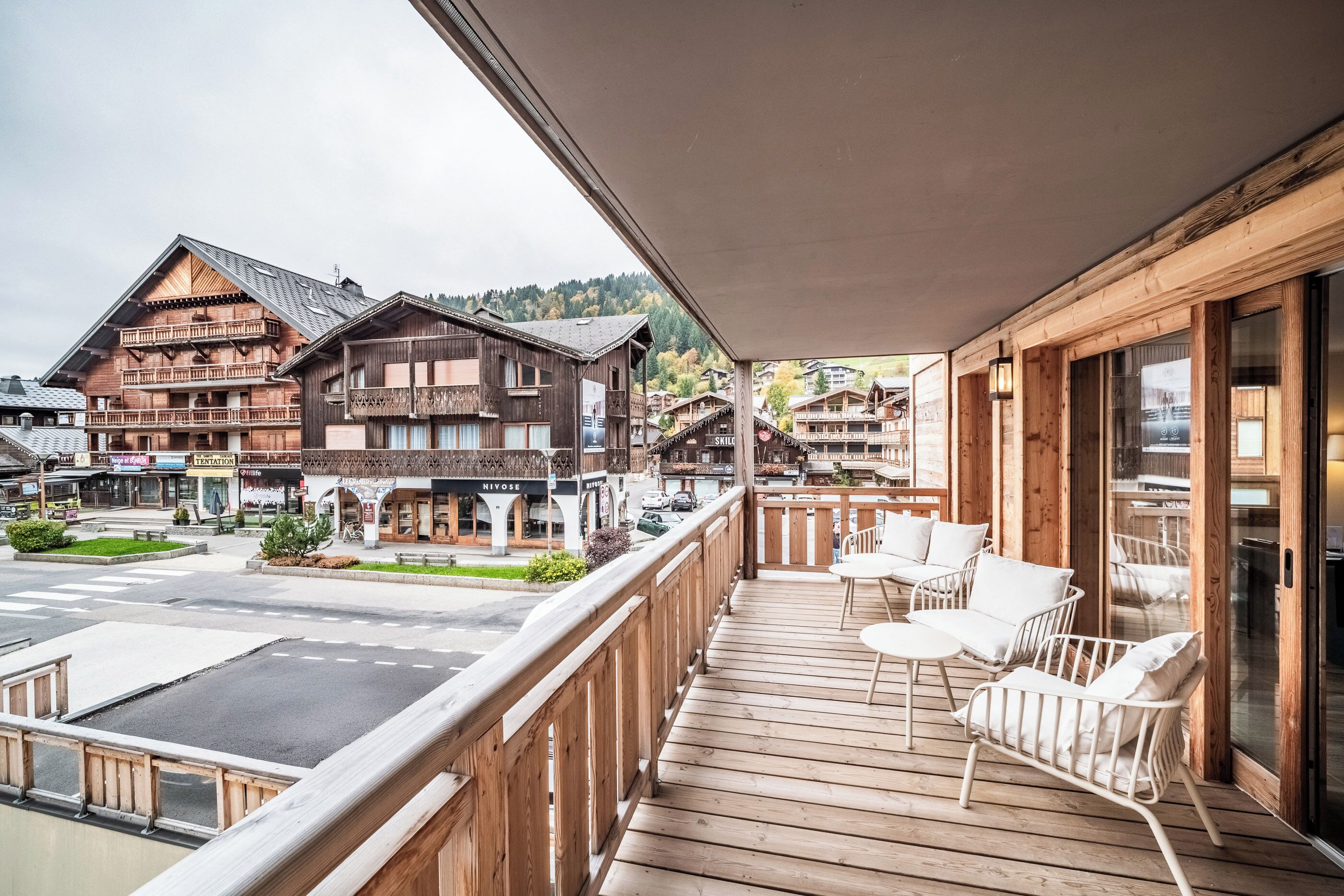 Een houten balkon met witte rieten meubels, met uitzicht op een straat met traditionele gebouwen en een helder zicht op de omgeving.