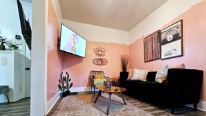 A living room with a black sofa, a wooden coffee table, a patterned rug, a cactus plant, and a TV mounted on the wall.