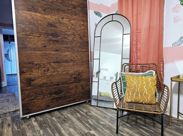 Une pièce avec un plancher en bois, une chaise en osier avec un coussin jaune, un miroir à motifs en arc de cercle et un rideau rouge.
