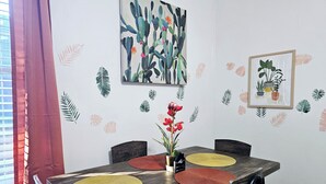 A dining area with a wooden table, two chairs, and a vase with red flowers. There are wall decals of leaves and a painting of cacti.