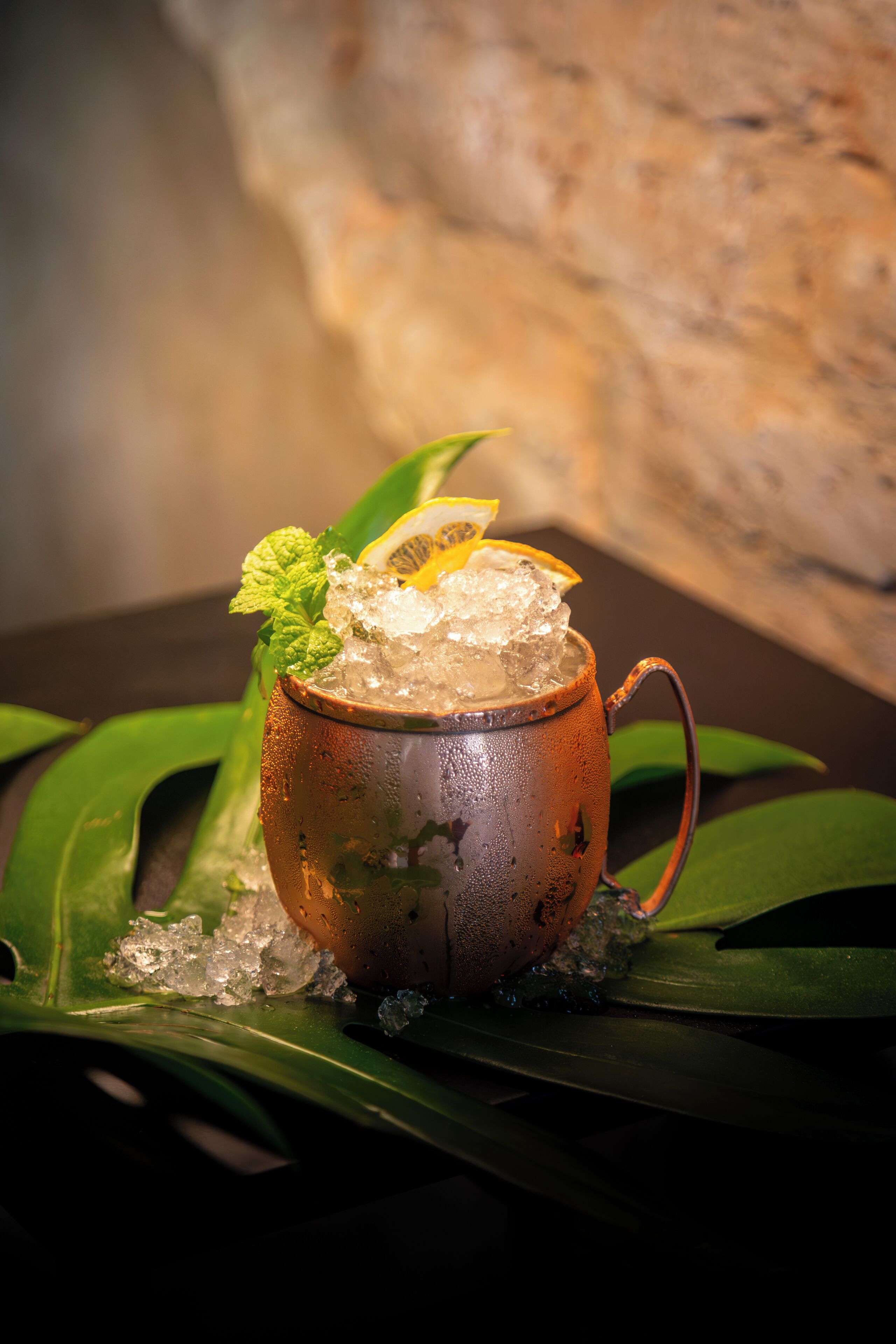 Un cocktail servi dans un mug rustique avec des glaçons, garni de menthe et d’agrumes, posé sur une surface sombre parsemée de feuilles vertes.