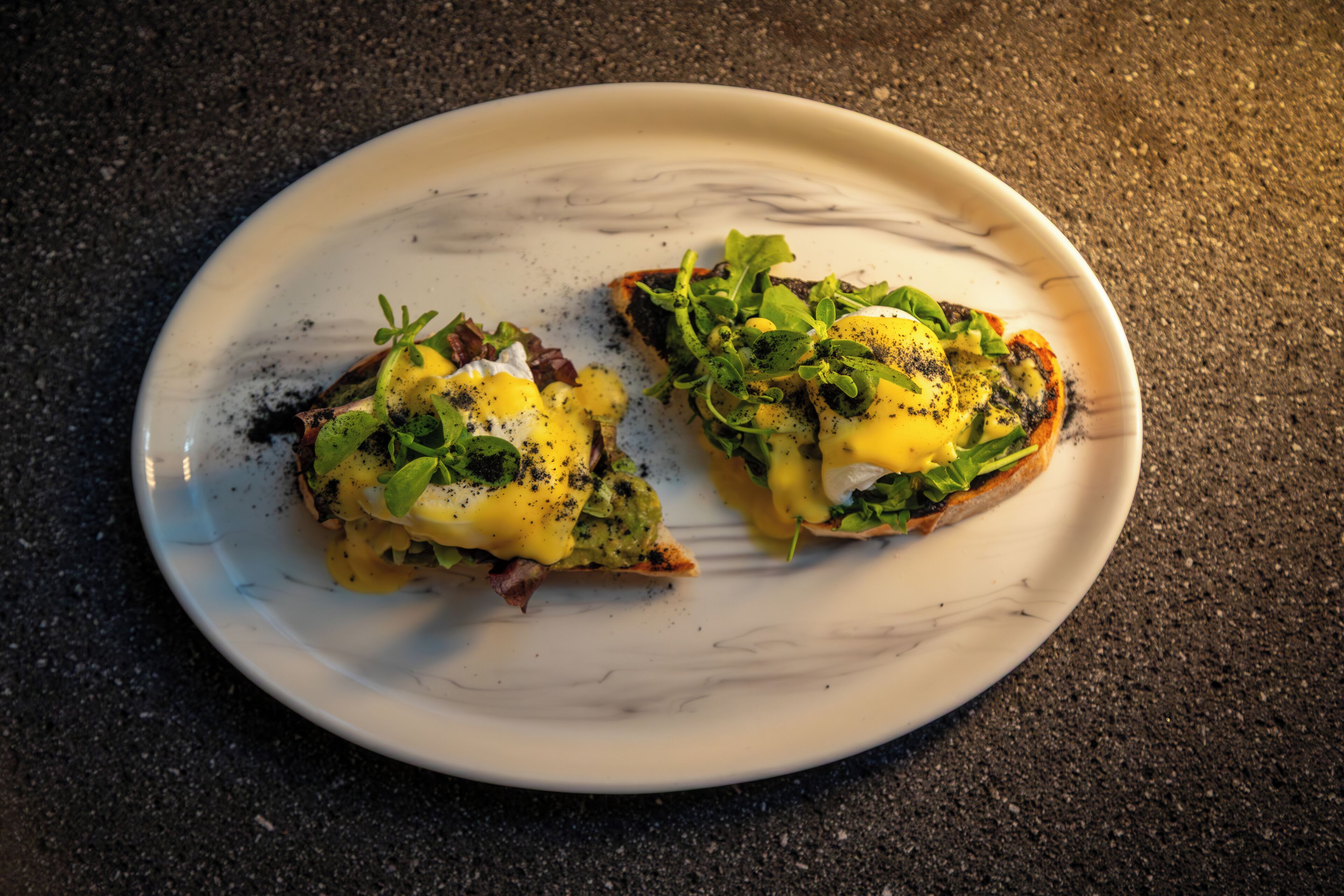 Deux sandwichs ouverts, présentés sur une assiette blanche à motifs marbrés, et garnis de verdure et d’une sauce jaune.