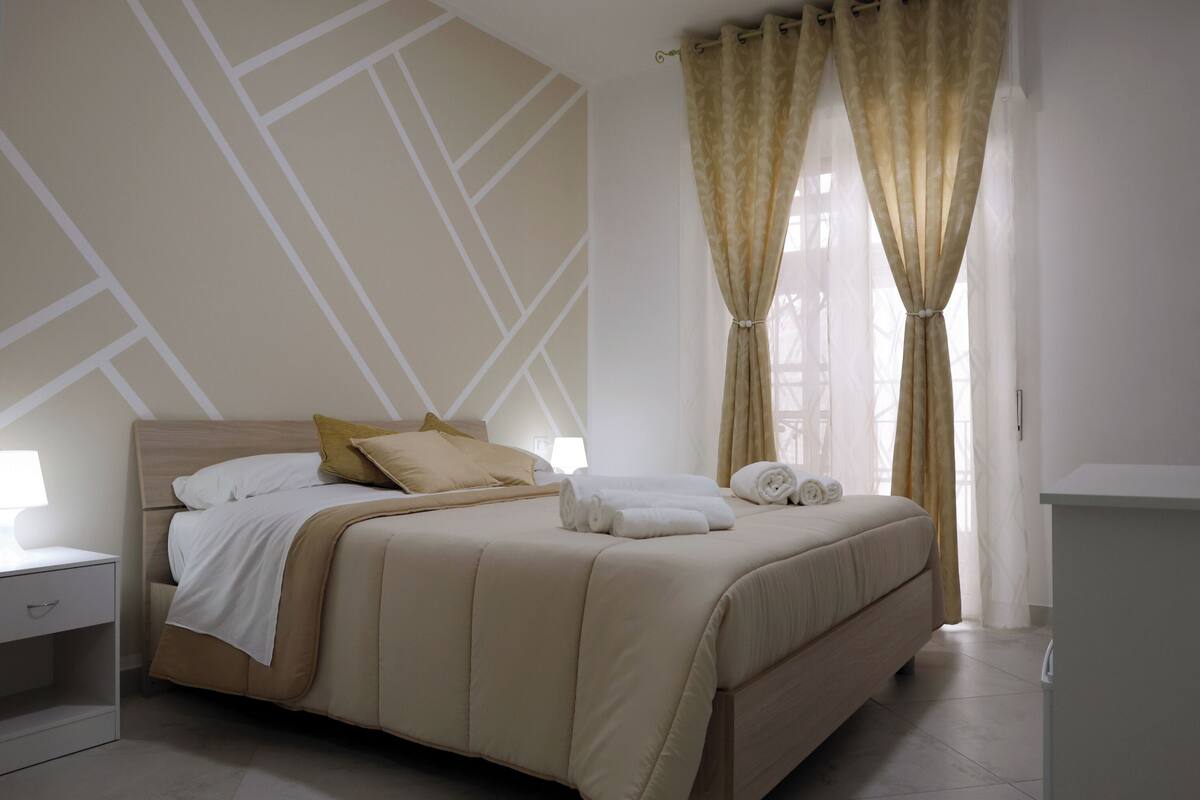 Una cama bien hecha con cabecera color beige, ropa de cama blanca y dos almohadas. Hay una mesita de noche con una lámpara junto a la cama.