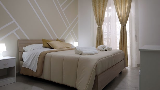 A neatly made bed with a beige headboard, white bedding, and two pillows. There is a nightstand with a lamp beside the bed.