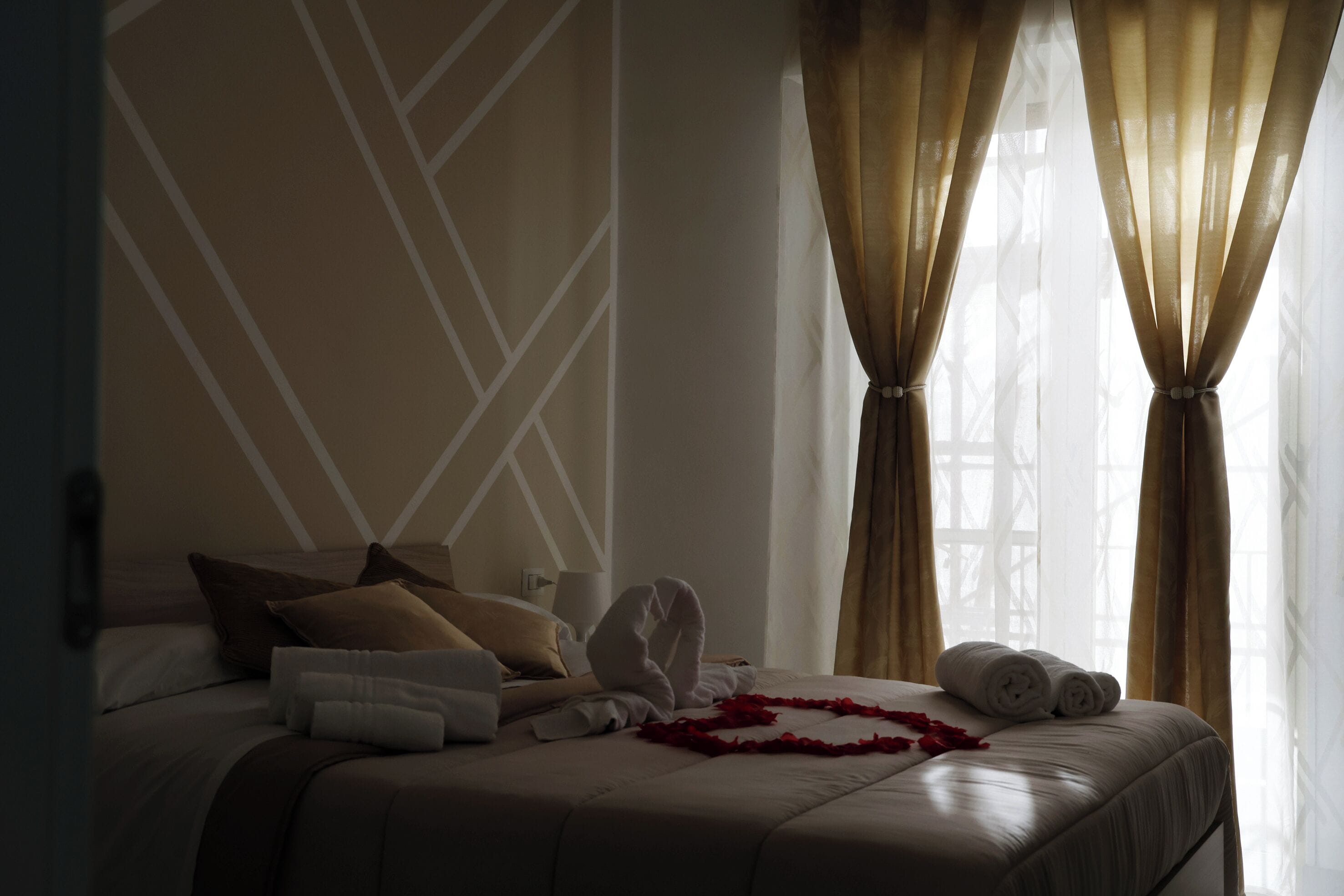 A hotel room with a neatly made bed, a decorative wall pattern, and a window with curtains.