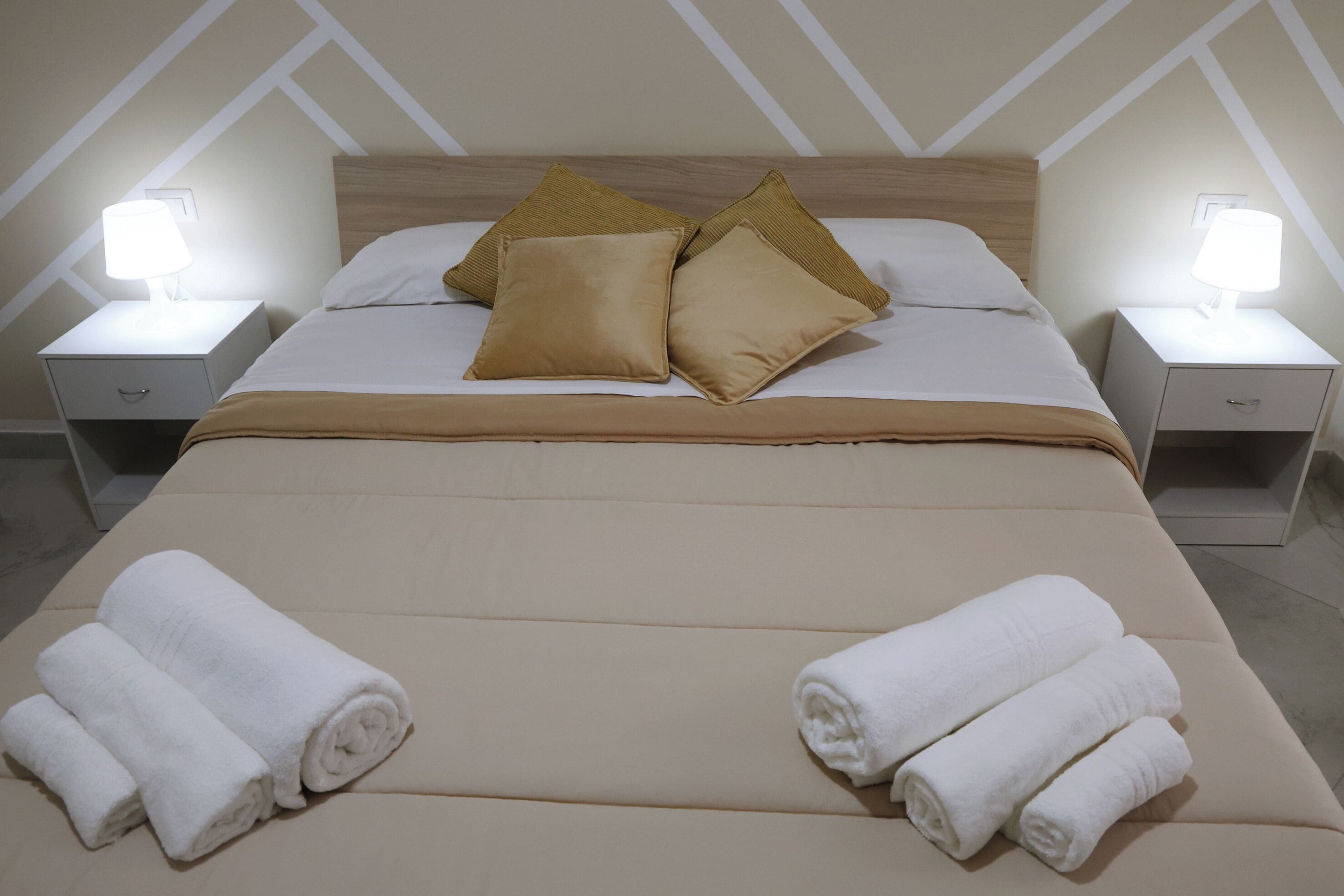 Una cama bien hecha con dos almohadas, dos mesitas de noche con lámparas y dos toallas enrolladas sobre la cama.