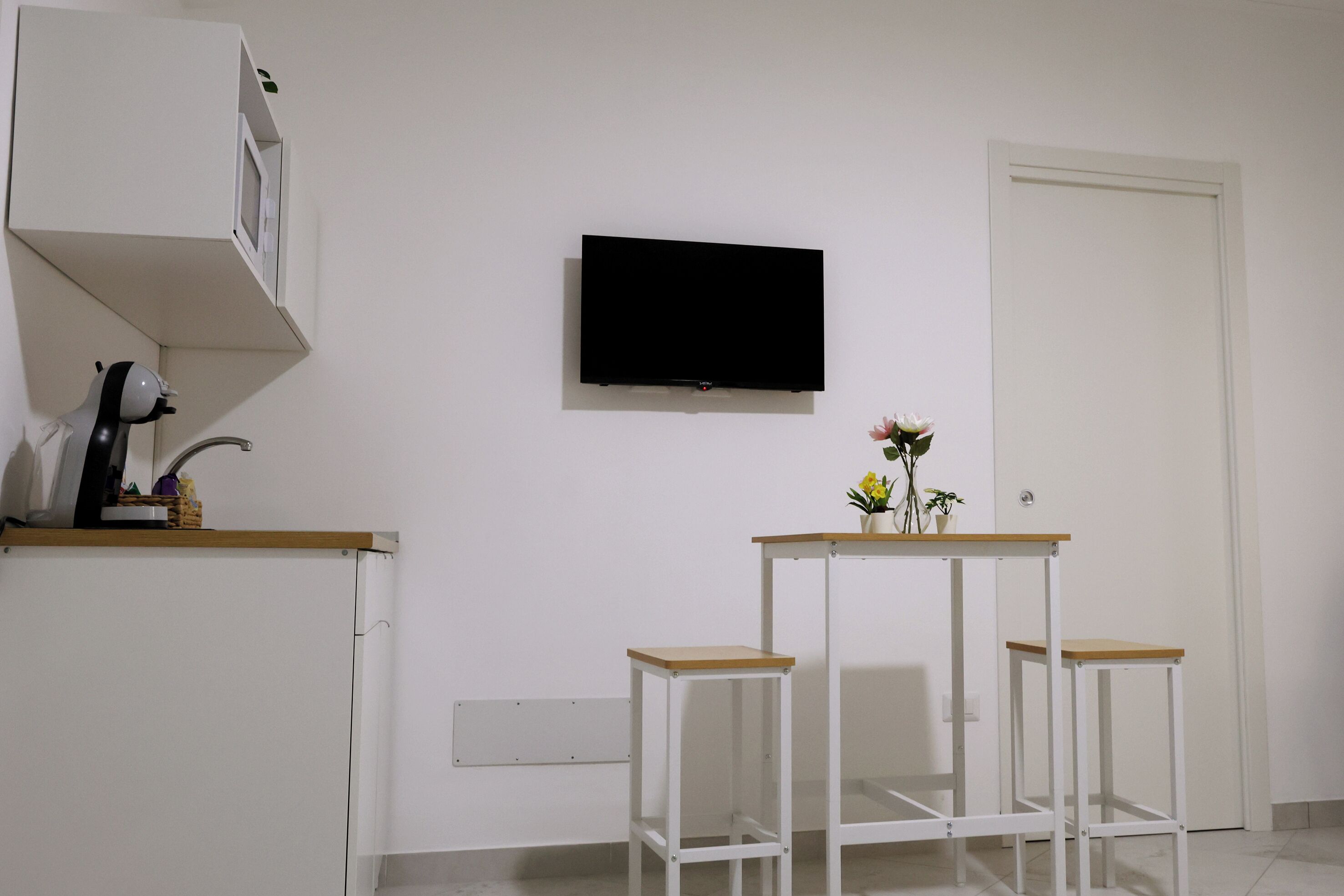Una habitación con pared blanca, un televisor de pantalla plana montado, una mesita con un florero y un mueble blanco con microondas.