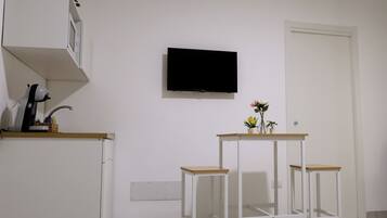 Una habitación con pared blanca, un televisor de pantalla plana montado, una mesita con un florero y un mueble blanco con microondas.