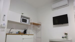 A compact kitchenette with a microwave, sink, and a small fridge. There is a white shelf above the sink with a plant, and a TV mounted on the wall.