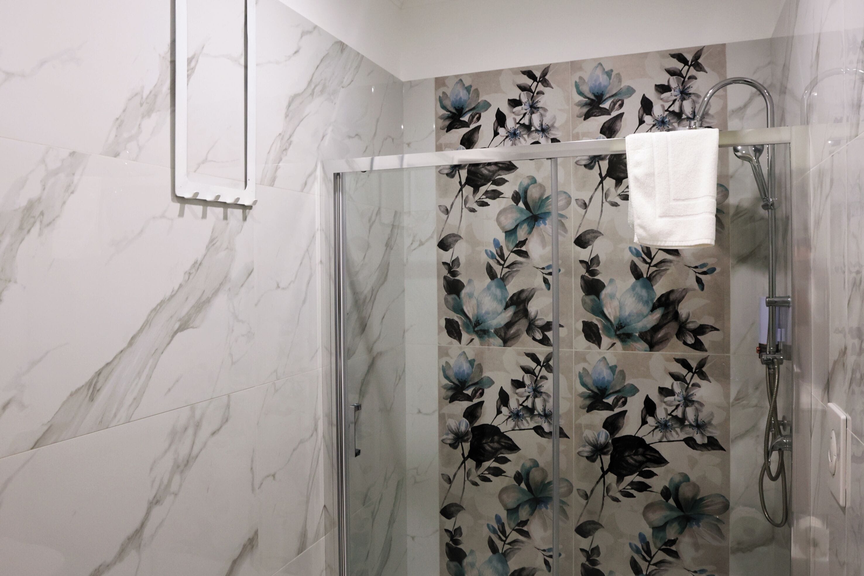 A glass-enclosed shower with a floral tile design, a white towel hanging on the door, and a marble floor.