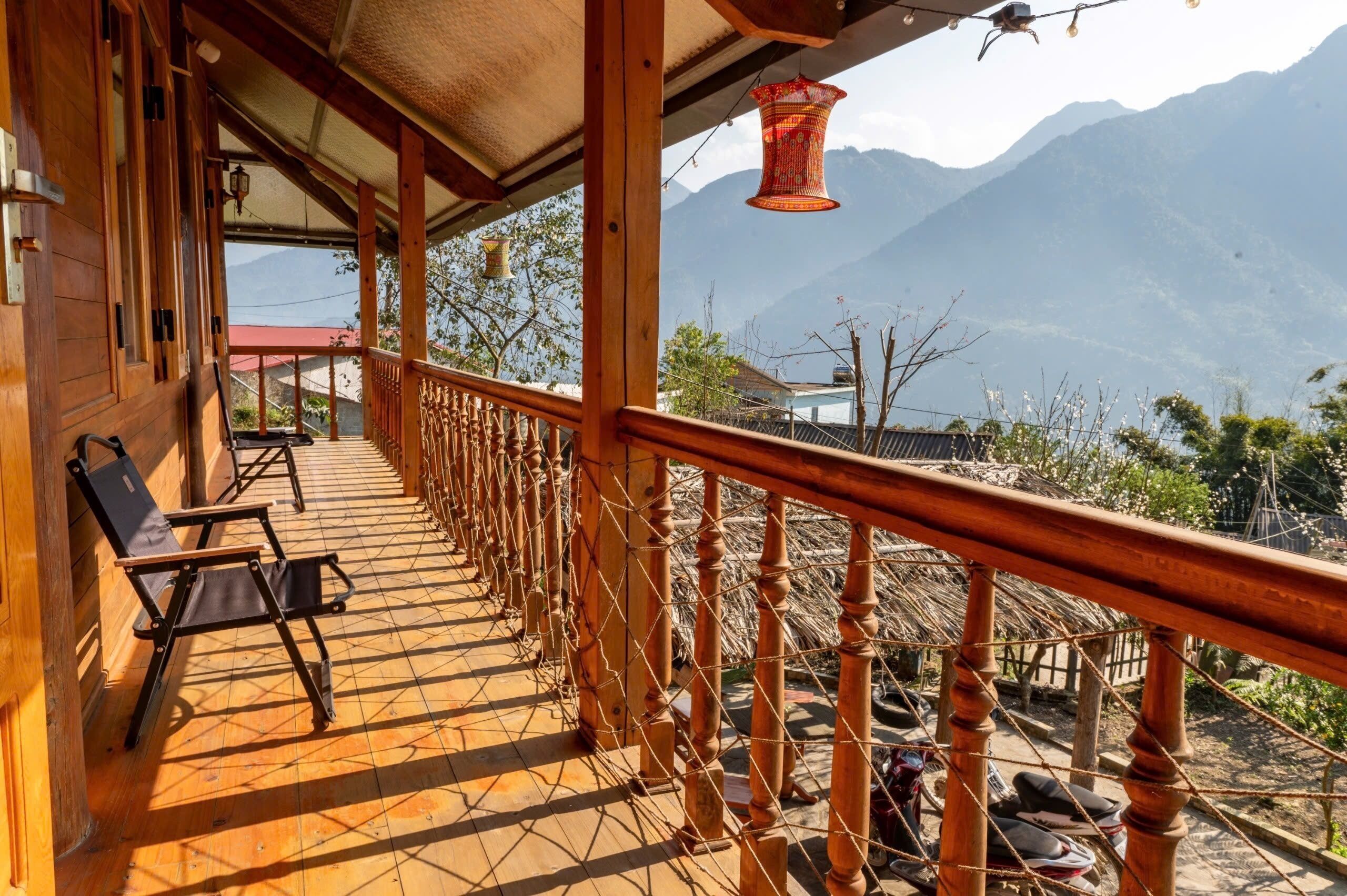 A wooden balcony with a view of mountains, a thatched roof structure, and a hanging lantern.