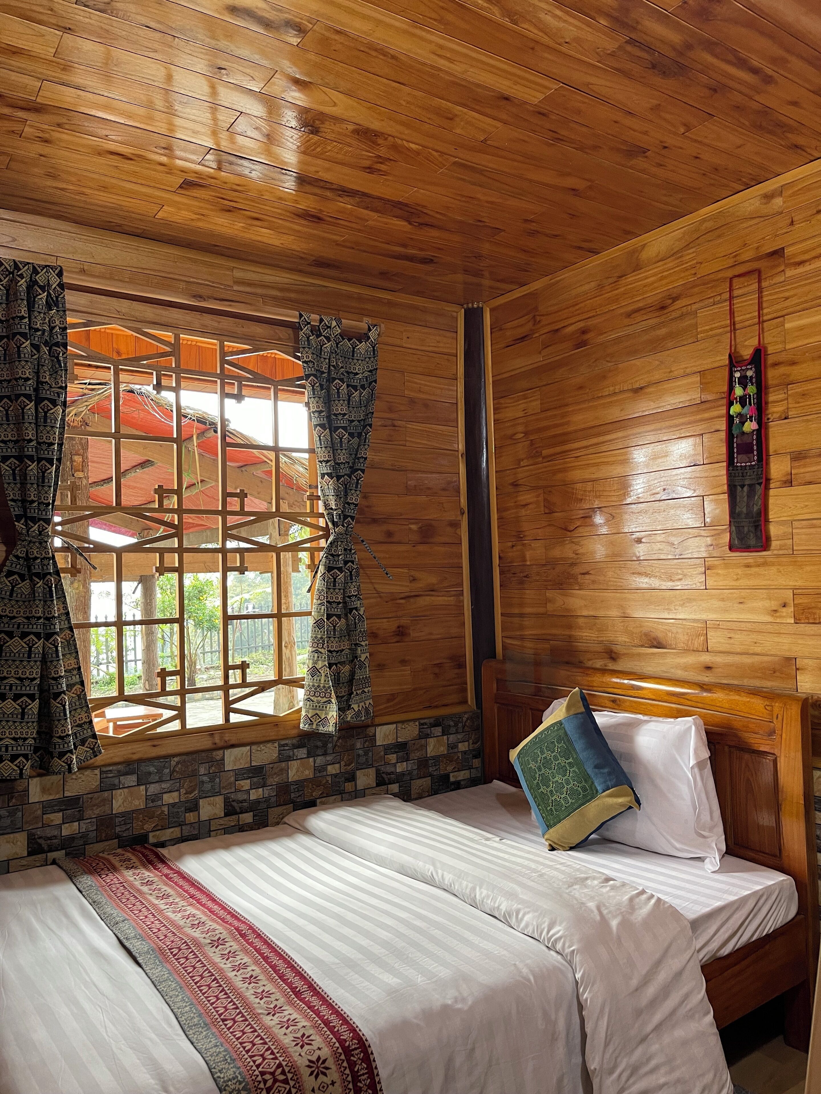 A wooden room with a bed, patterned curtains, and a view through a window.