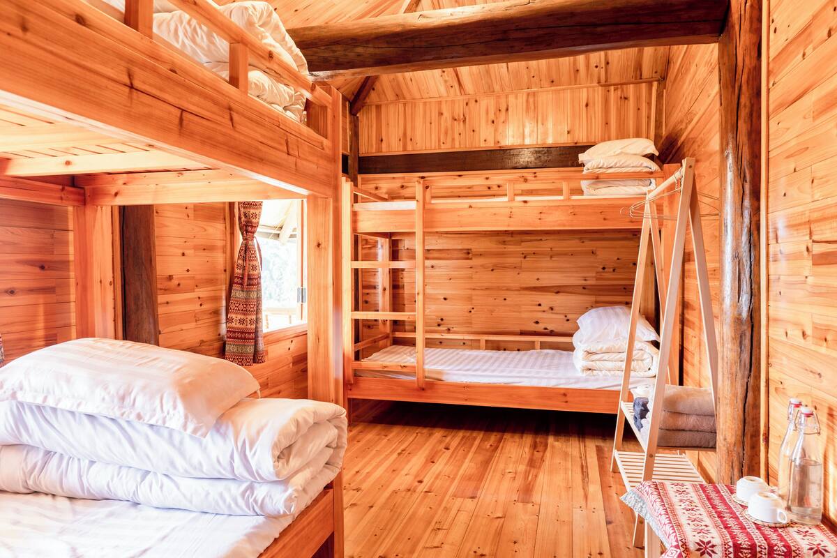 A wooden cabin with bunk beds, a small table with cups and a bottle, and a window with curtains.