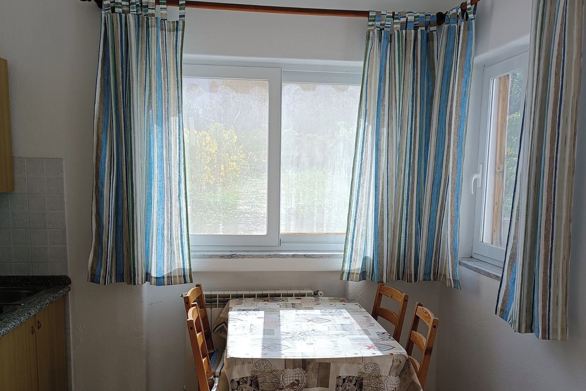 A dining area with a table and chairs, a window with curtains, and a kitchen area with a sink and cabinets.