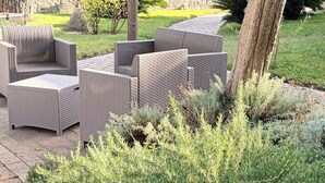 Wicker outdoor furniture arranged on a paved patio with a lush green shrubbery in the foreground. - The Vineyard (Forio)