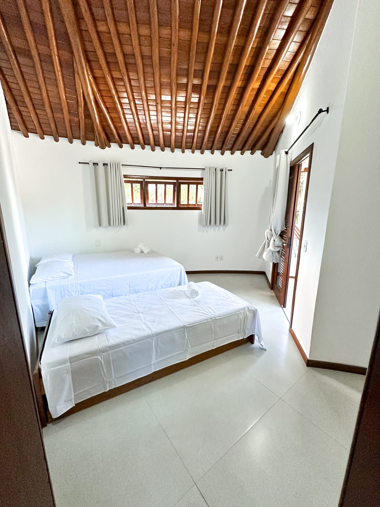 Une chambre avec un plafond en bois, un lit double, une fenêtre avec des rideaux et une porte dotée d’un porte-serviettes.