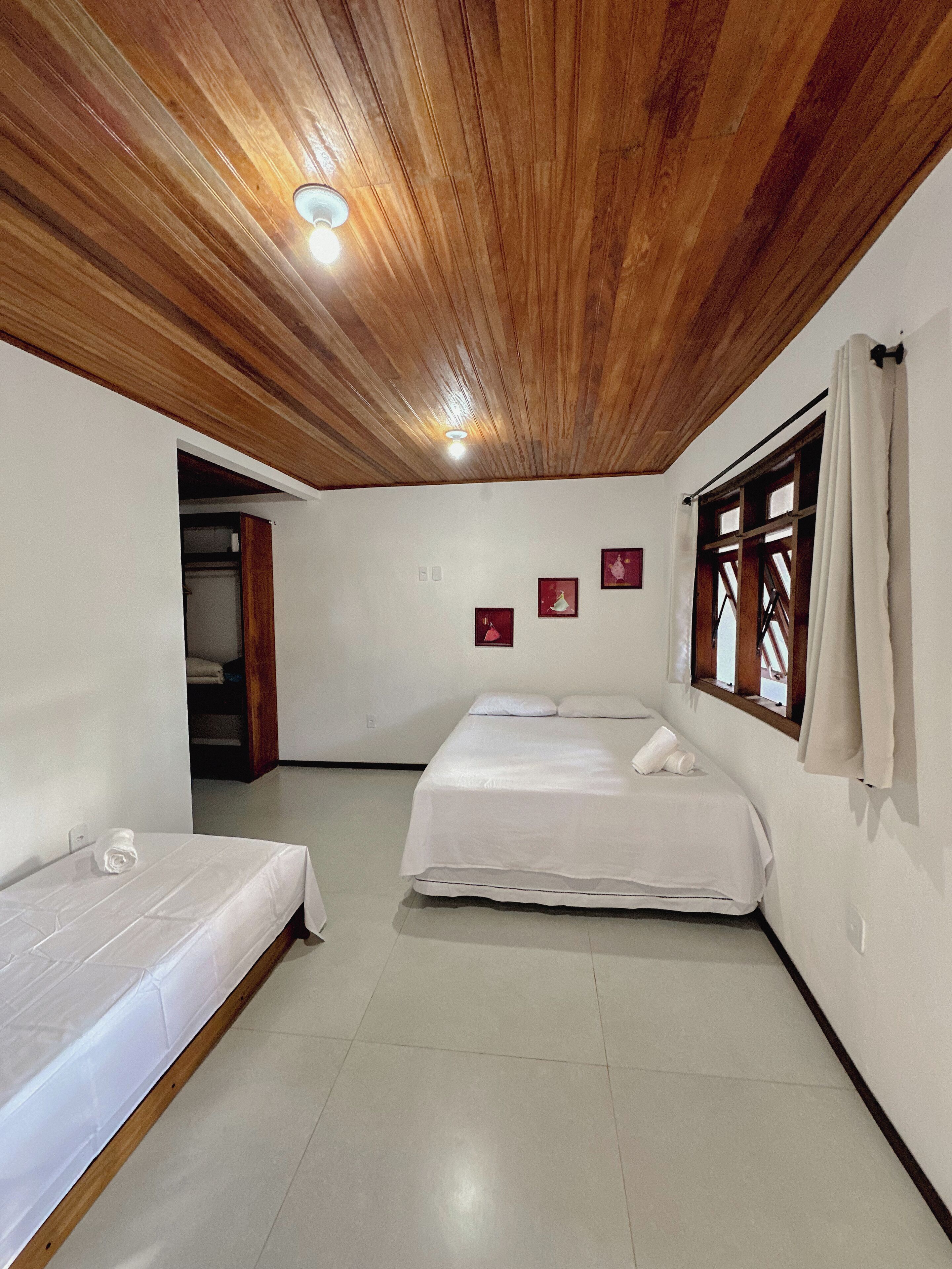 A room with two beds, wooden ceiling, white walls, and a window with curtains.