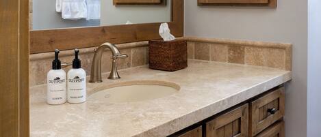 A bathroom with a large mirror, wooden cabinets, a sink, and two bottles of Outpost hand soap.