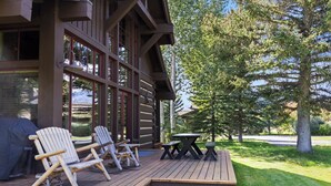 Una terraza de madera con sillas Adirondack y una mesa de picnic, con vista a un área cubierta de césped y árboles.