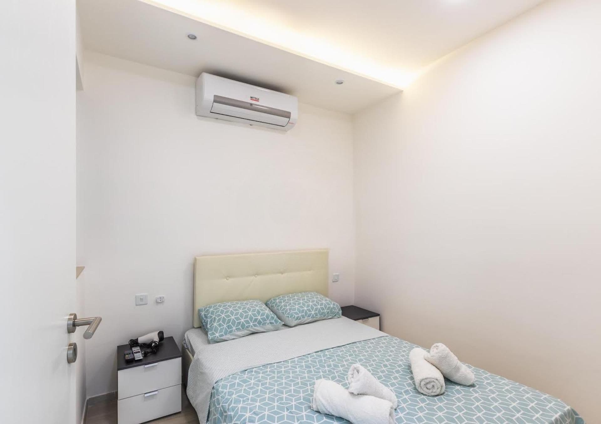 Una habitación de hotel con una cama, mesitas de noche, un equipo de aire acondicionado y una toalla sobre la cama.