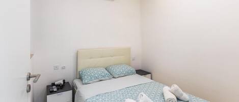 Una habitación de hotel con una cama, mesitas de noche, un equipo de aire acondicionado y una toalla sobre la cama.