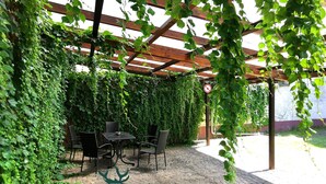 Un área de estar al aire libre cubierta con una pérgola, rodeada de frondosas enredaderas verdes y un piso de adoquinado.