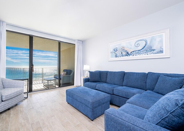 Una sala de estar moderna con un sofá azul, un sillón pequeño y un ventanal de vidrio que da a un balcón con vistas al mar.
