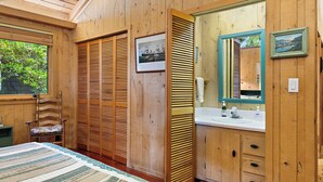 Una habitación de madera con una cama, una silla, un ventanal grande, un espejo y un lavamanos.