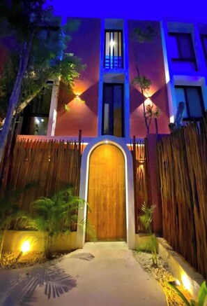 Enter through the welcoming wooden door, flanked by a bamboo fence, into a building with a spacious balcony and railing.