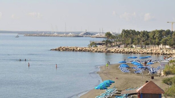 Una playa con un rompeolas rocoso, barcos y una fila de sombrillas azules.