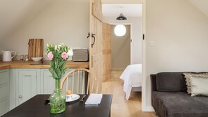 A cozy kitchen with a dining table, a vase with flowers, and a vintage radio. A wooden door leads to a bedroom with a bed and a round ceiling light.