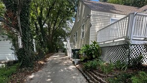 A narrow concrete pathway flanked by lush greenery and white wooden houses with visible windows and a white fence.