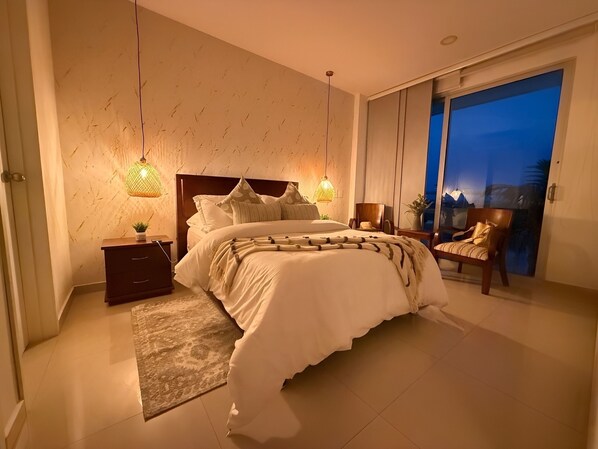 Ein Schlafzimmer mit einem großen Bett, Nachttischen, einem Beistelltisch, einem Sessel und Blick ins Freie.