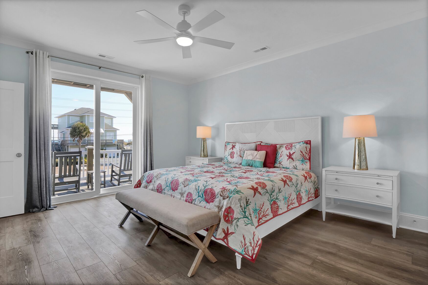 A bedroom with a bed, two bedside tables, a bench, a ceiling fan, and a view through a sliding glass door to the outside.